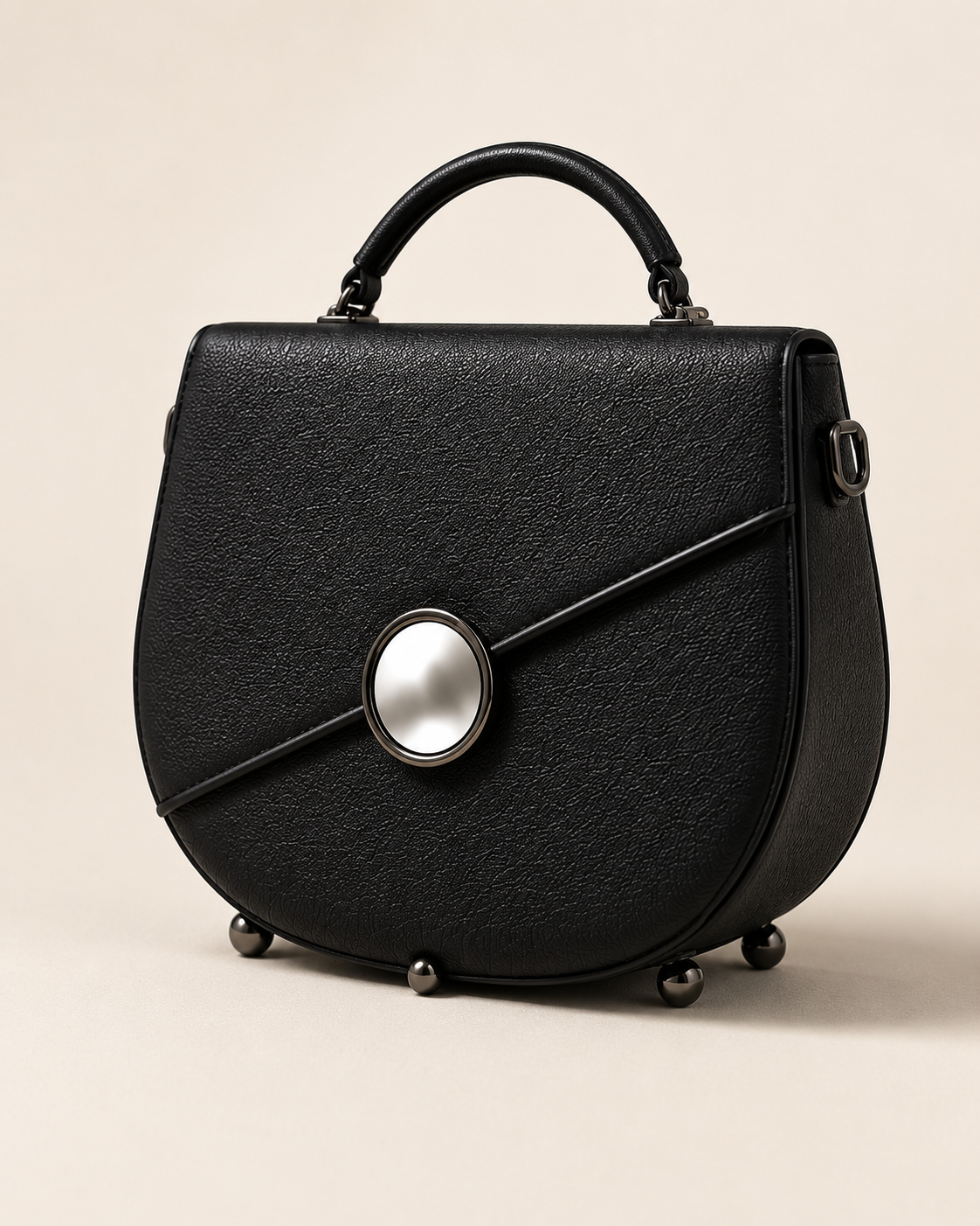 Rosie handbag in Noir — rounded saddle silhouette in black pebbled full-grain leather, sculpted top handle, diagonal seam closed by a circular mirror clasp in a gunmetal bezel, small gunmetal ball feet